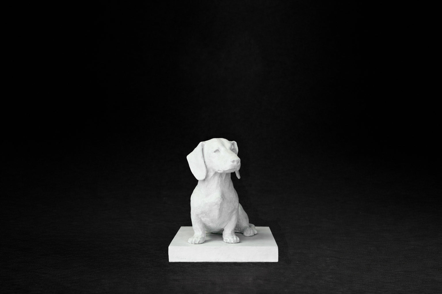 White plaster sculpture of a seated dachshund on a rectangular base, gazing forward.