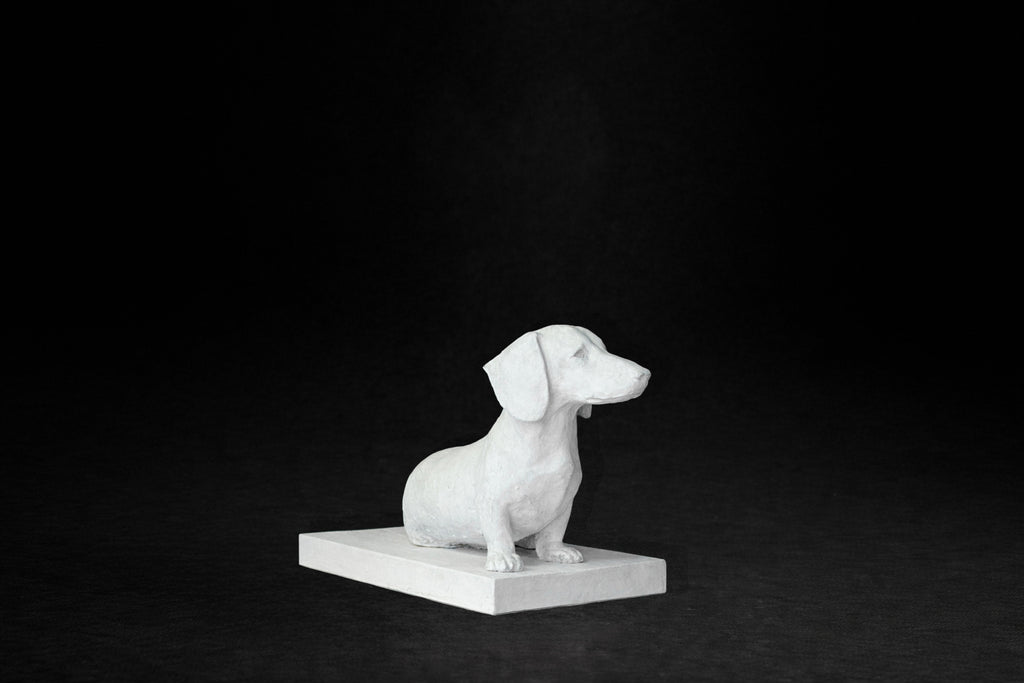 White plaster sculpture of a seated dachshund on a rectangular base, gazing forward.