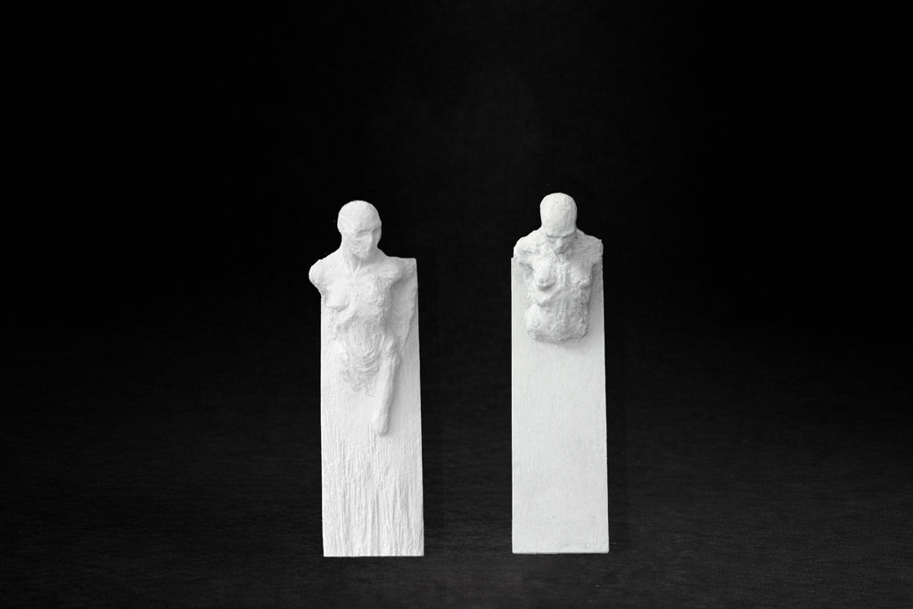 Two white plaster wall-mounted figural panels depicting elongated human forms emerging from vertical plaques.