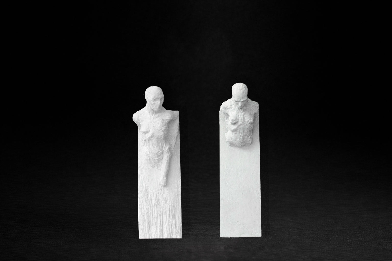 Two white plaster wall-mounted figural panels depicting elongated human forms emerging from vertical plaques.