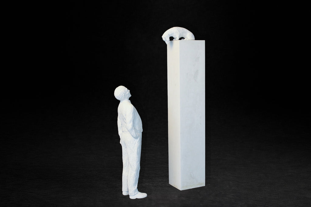 White plaster sculpture of a man standing beside a tall rectangular plinth, looking up at a dachshund peering over the edge.