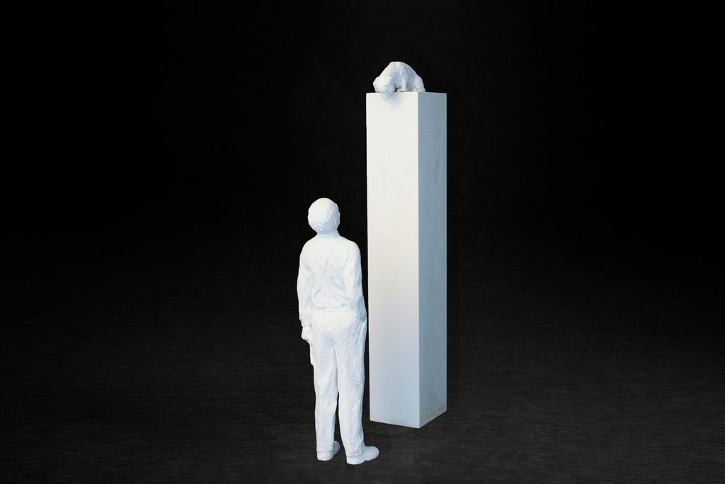 White plaster sculpture of a man standing beside a tall rectangular plinth, looking up at a dachshund peering over the edge.