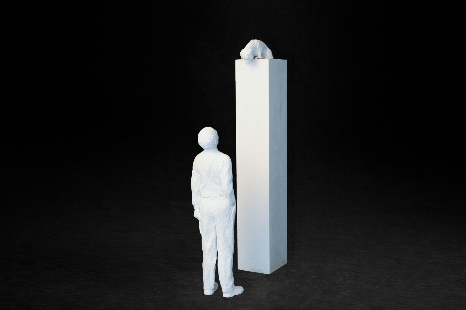 White plaster sculpture of a man standing beside a tall rectangular plinth, looking up at a dachshund peering over the edge.