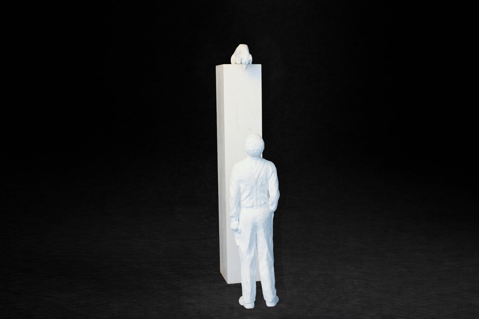 White plaster sculpture of a man standing beside a tall rectangular plinth, looking up at a dachshund peering over the edge.