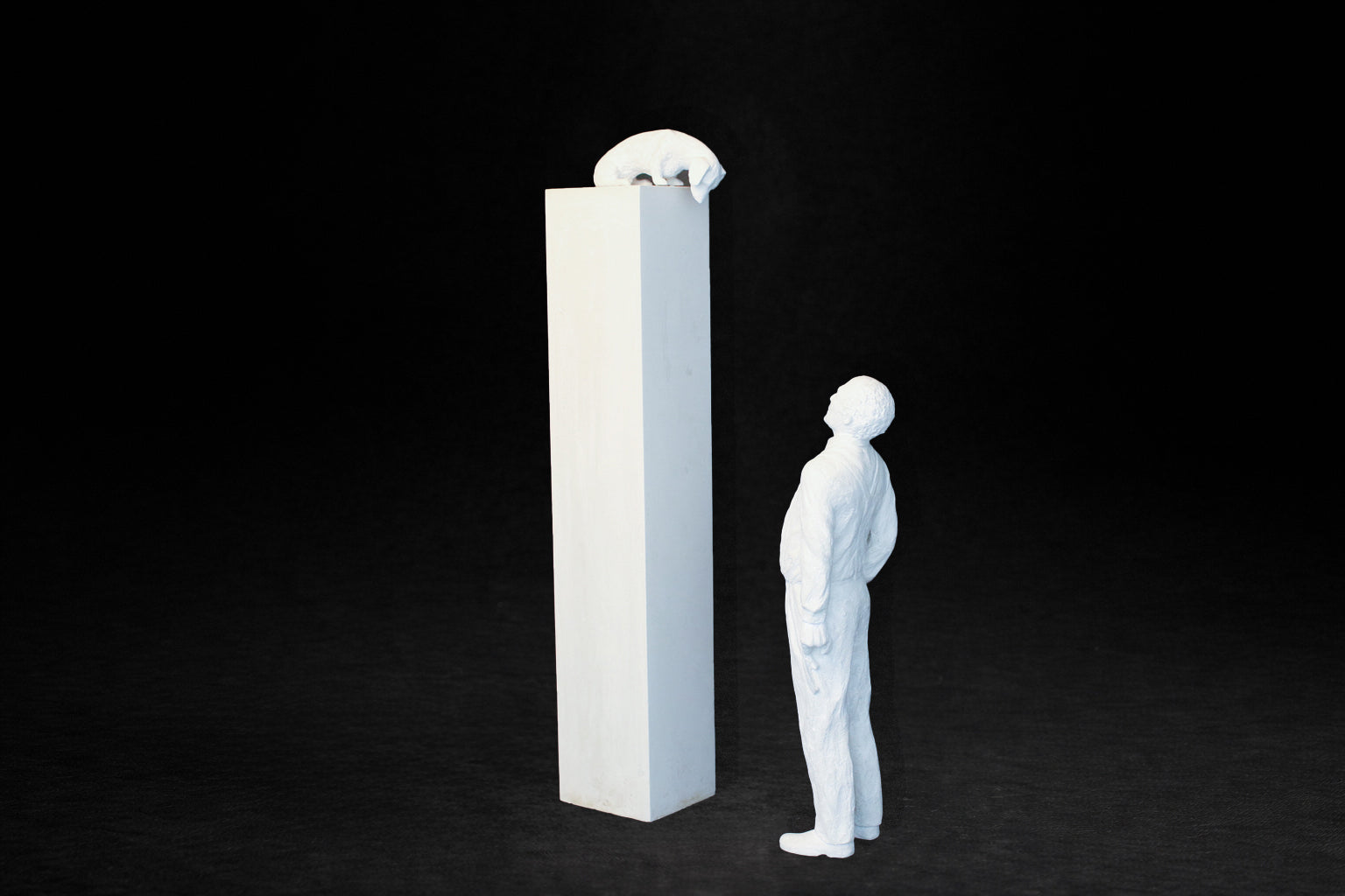 White plaster sculpture of a man standing beside a tall rectangular plinth, looking up at a dachshund peering over the edge.