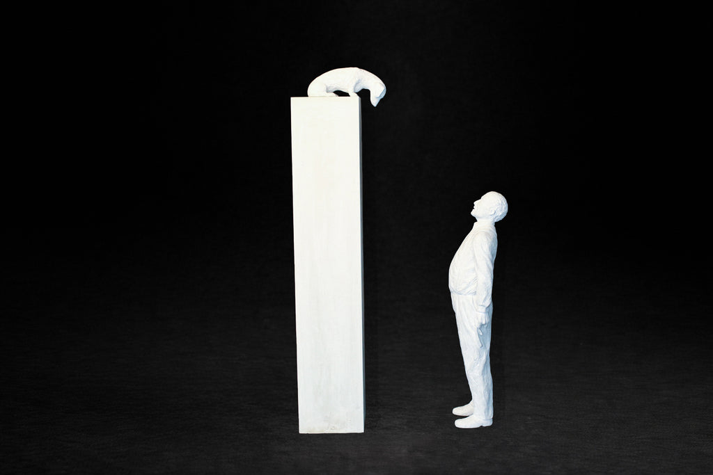 White plaster sculpture of a man standing beside a tall rectangular plinth, looking up at a dachshund peering over the edge.