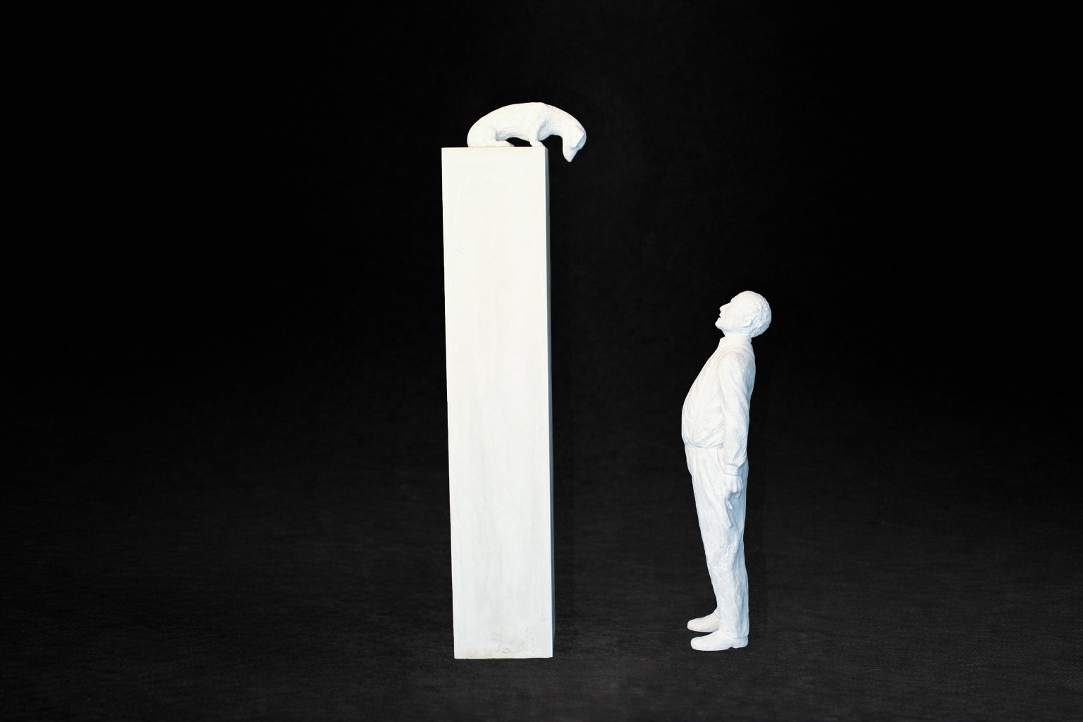 White plaster sculpture of a man standing beside a tall rectangular plinth, looking up at a dachshund peering over the edge.