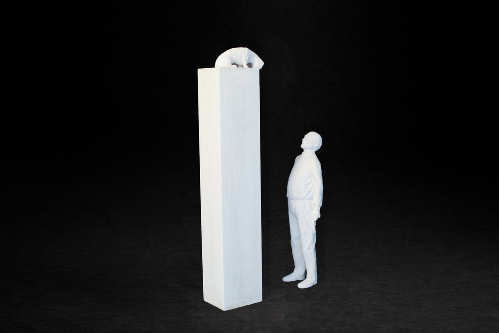 White plaster sculpture of a man standing beside a tall rectangular plinth, looking up at a dachshund peering over the edge.