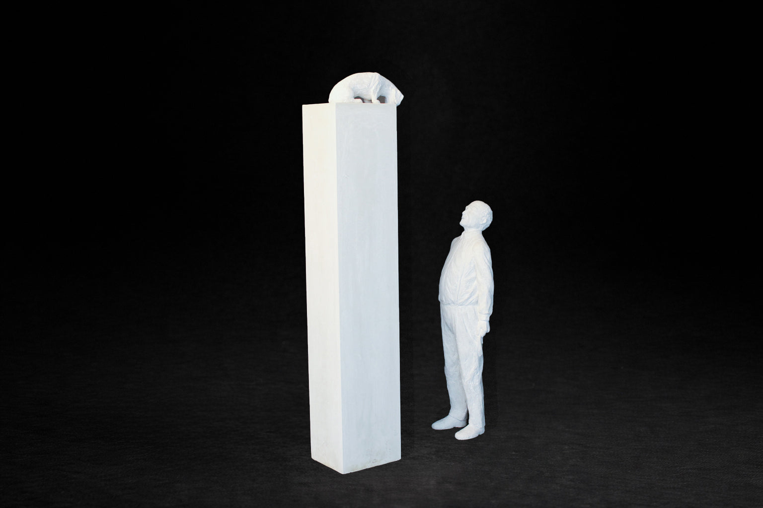 White plaster sculpture of a man standing beside a tall rectangular plinth, looking up at a dachshund peering over the edge.