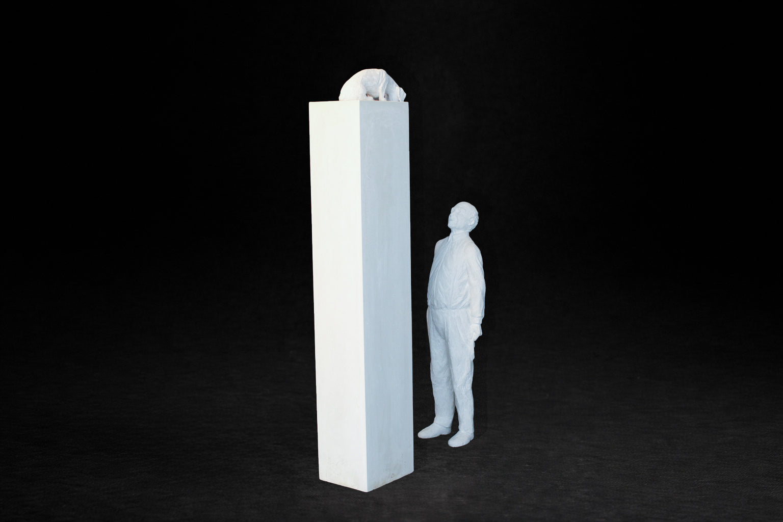 White plaster sculpture of a man standing beside a tall rectangular plinth, looking up at a dachshund peering over the edge.