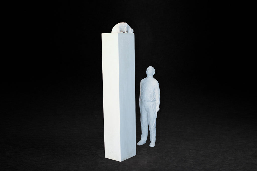 White plaster sculpture of a man standing beside a tall rectangular plinth, looking up at a dachshund peering over the edge.