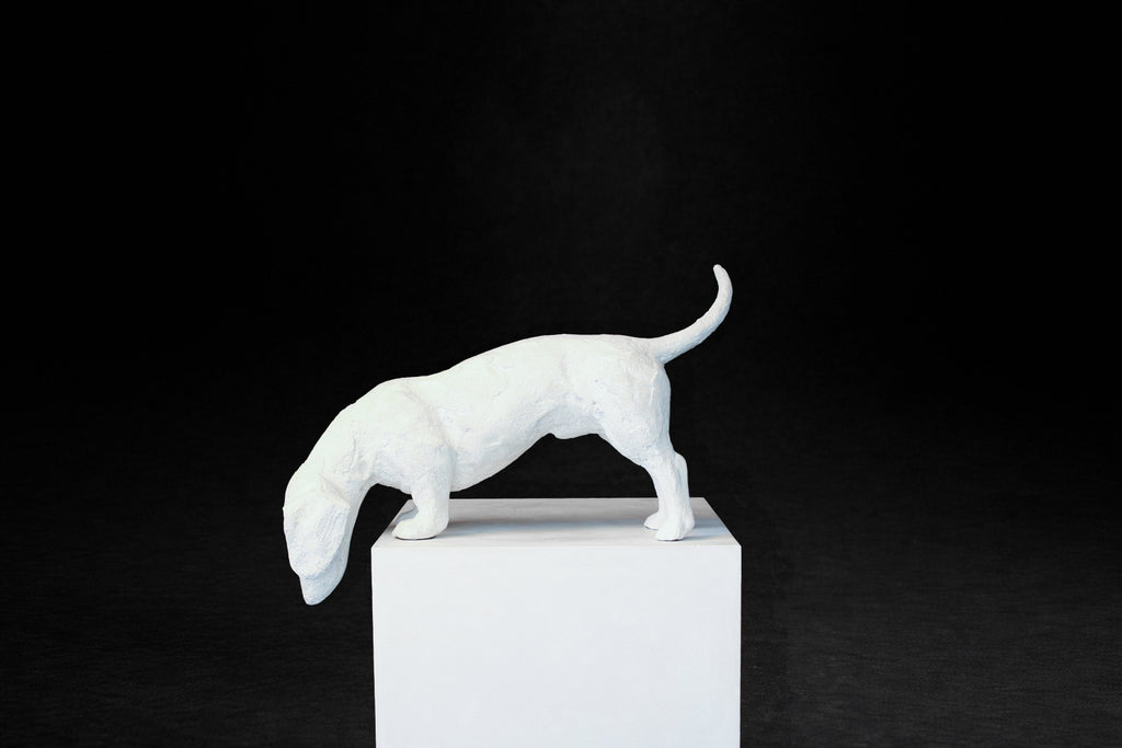 White plaster sculpture of a dachshund leaning forward off the edge of a square plinth.
