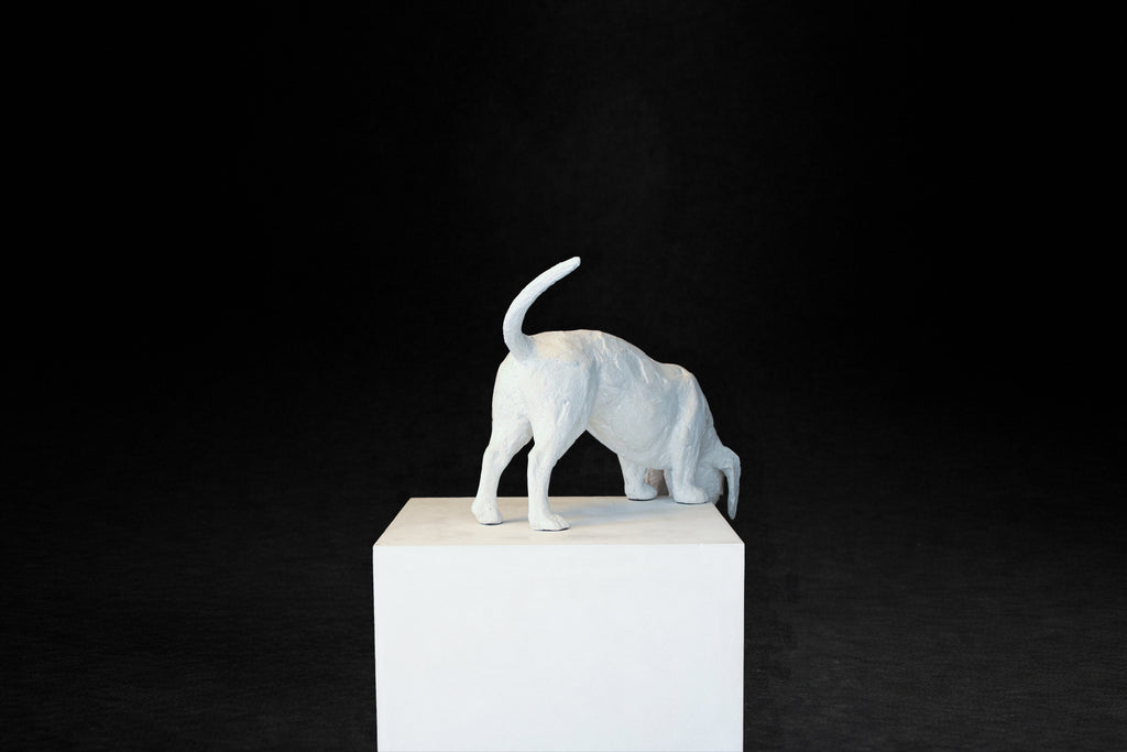 White plaster sculpture of a dachshund leaning forward off the edge of a square plinth.