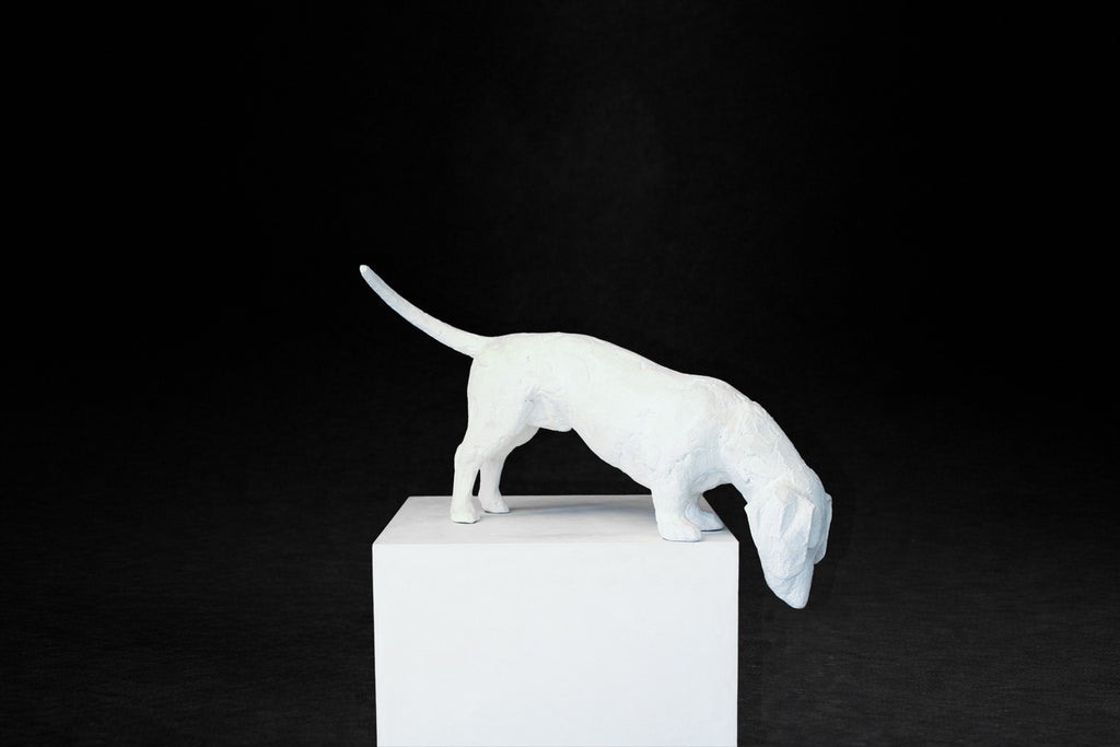 White plaster sculpture of a dachshund leaning forward off the edge of a square plinth.