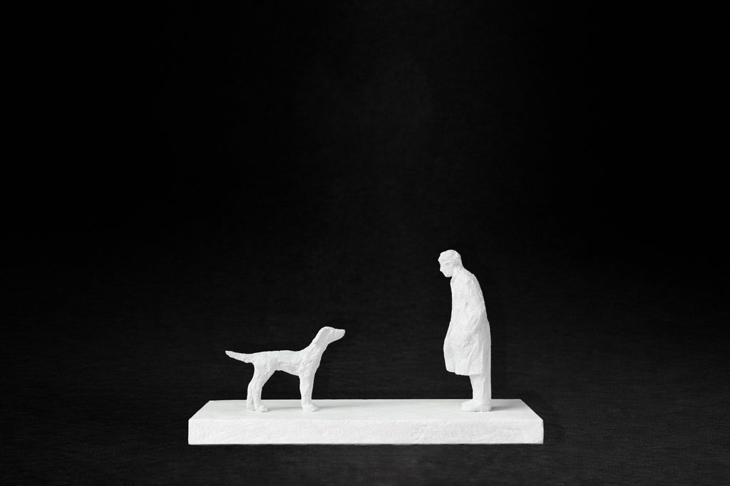 White plaster sculpture of a person and a dog facing each other on a rectangular base.