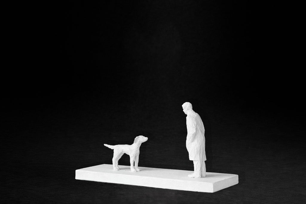 White plaster sculpture of a person and a dog facing each other on a rectangular base.