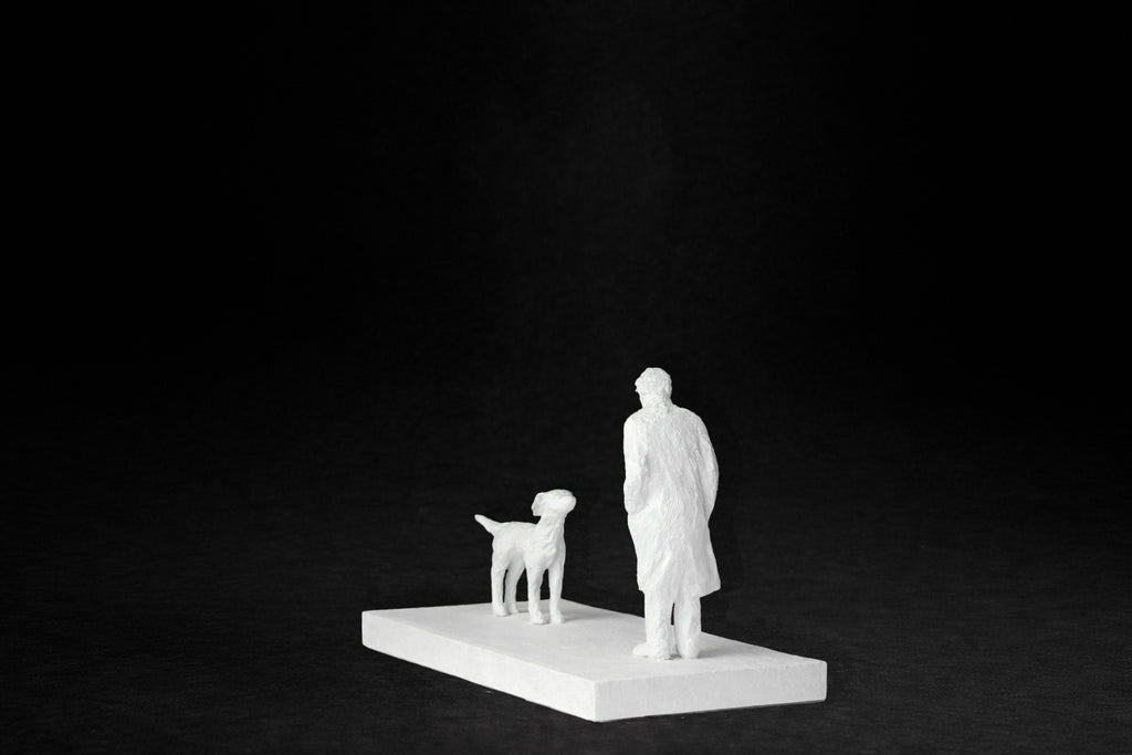 White plaster sculpture of a person and a dog facing each other on a rectangular base.