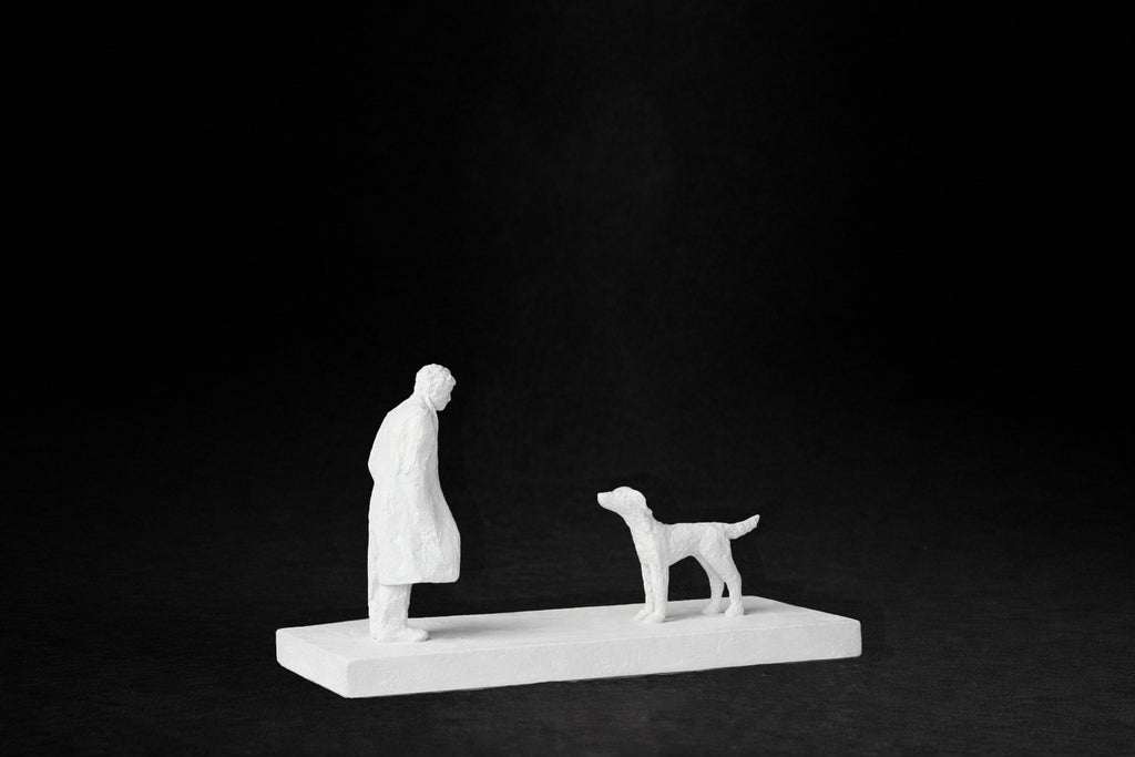White plaster sculpture of a person and a dog facing each other on a rectangular base.