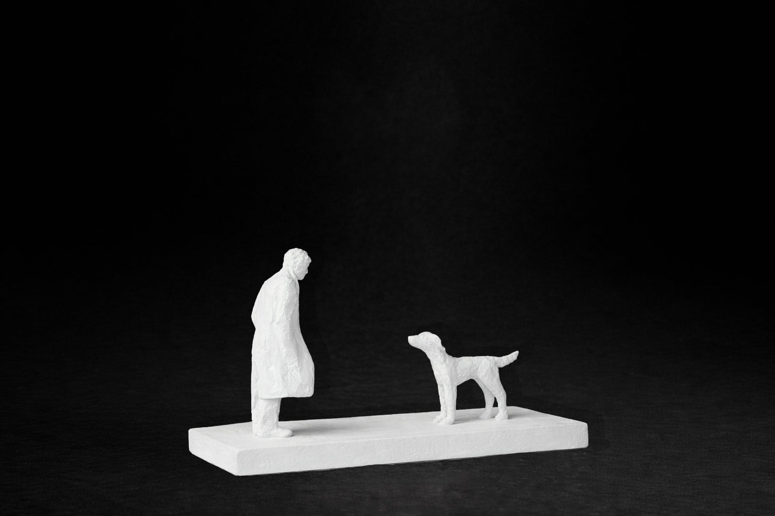 White plaster sculpture of a person and a dog facing each other on a rectangular base.