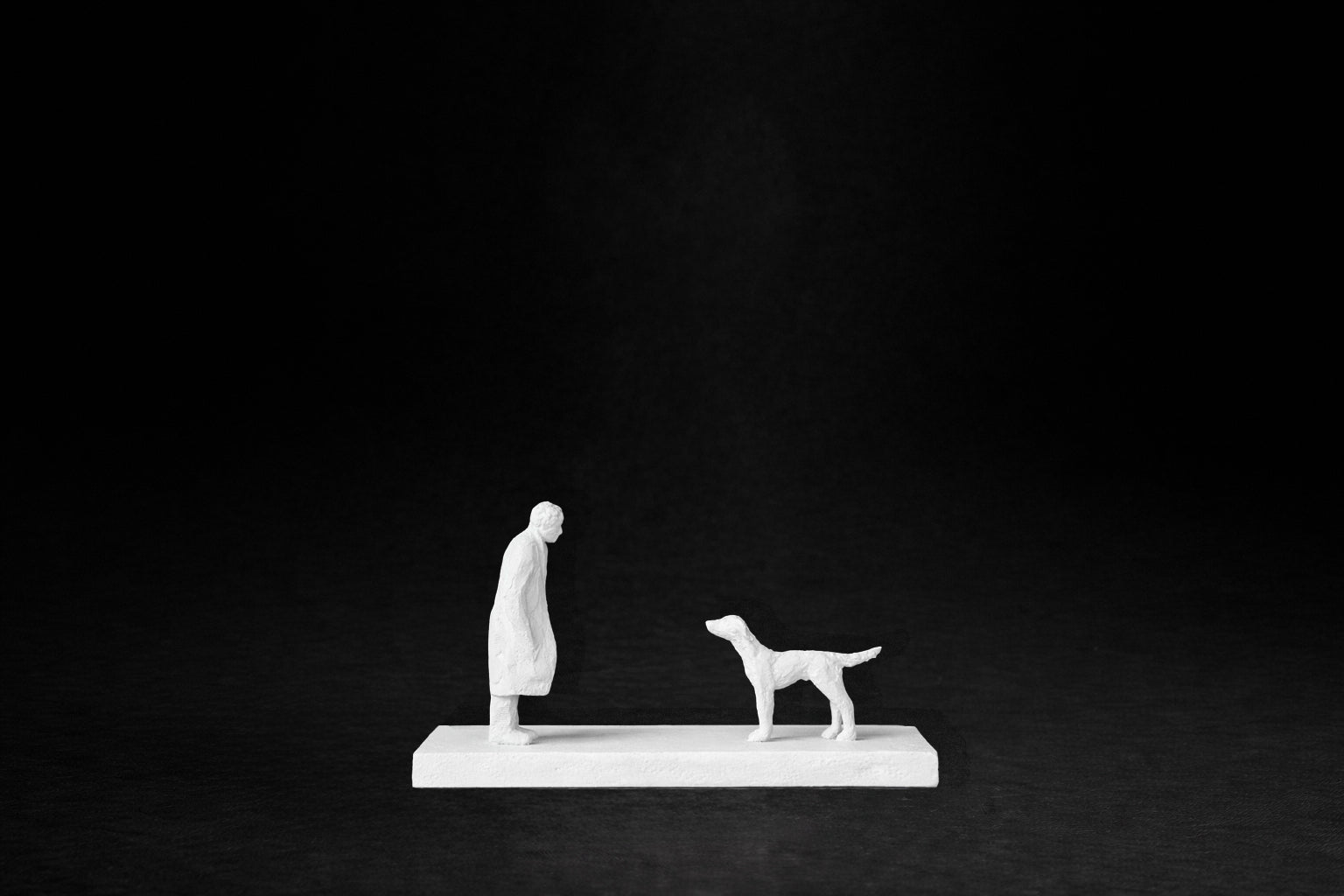 White plaster sculpture of a person and a dog facing each other on a rectangular base.