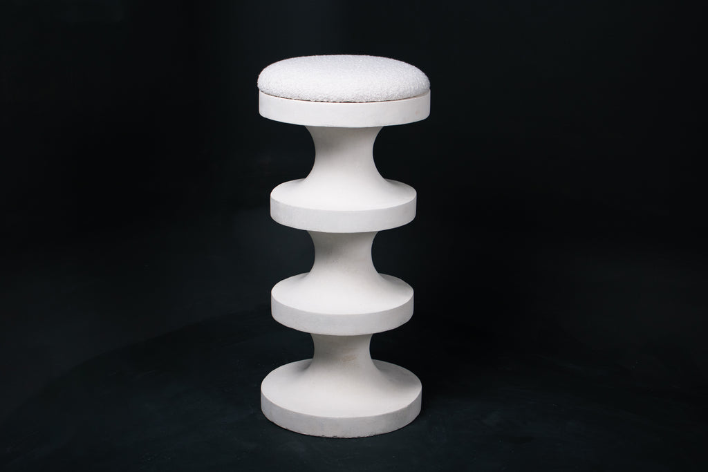 The Tambour barstool in white. The sculptural pedestal base features three stacked hourglass tiers that widen and narrow in alternating rhythm, topped with a round white bouclé upholstered seat.