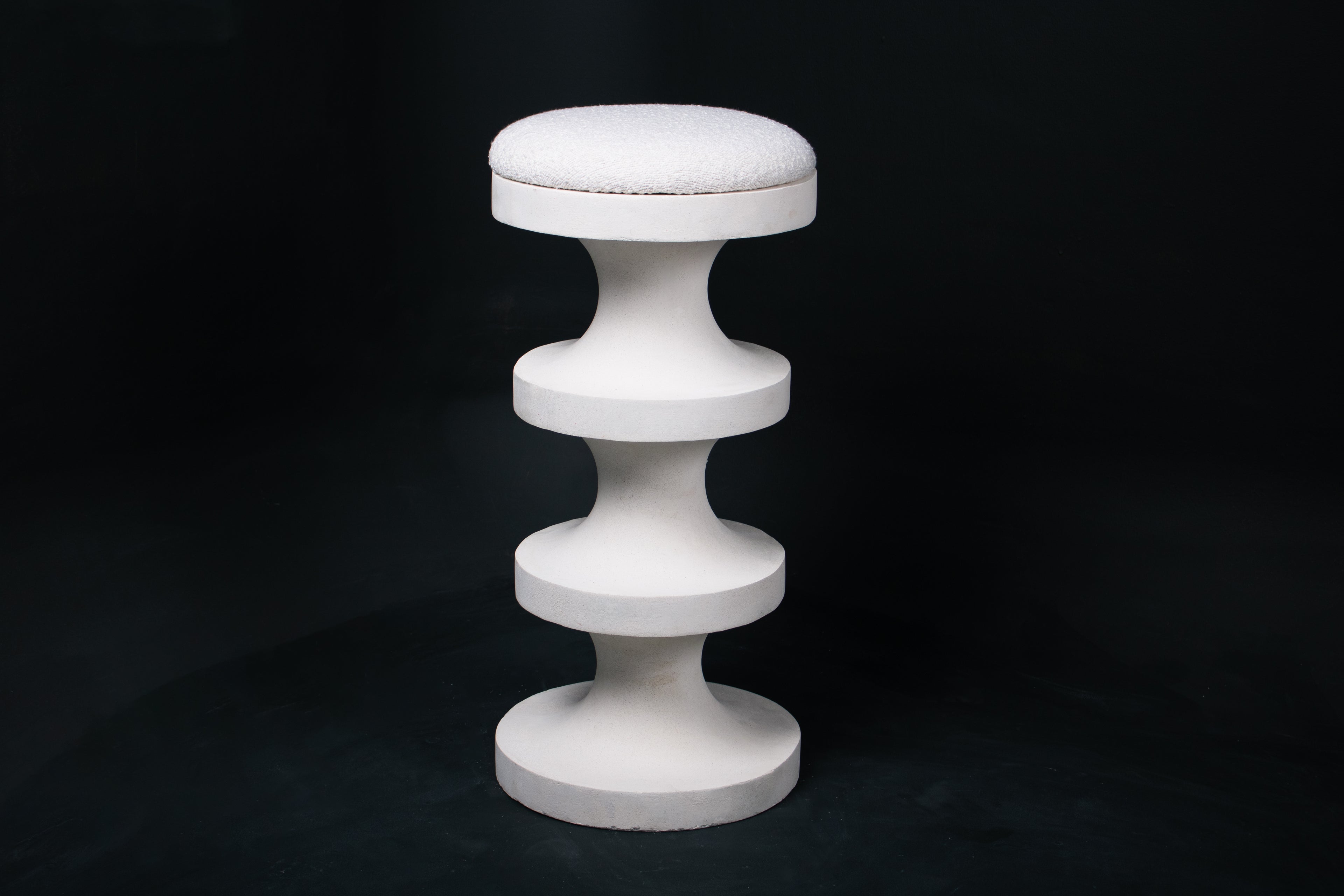 The Tambour barstool in white. The sculptural pedestal base features three stacked hourglass tiers that widen and narrow in alternating rhythm, topped with a round white bouclé upholstered seat.