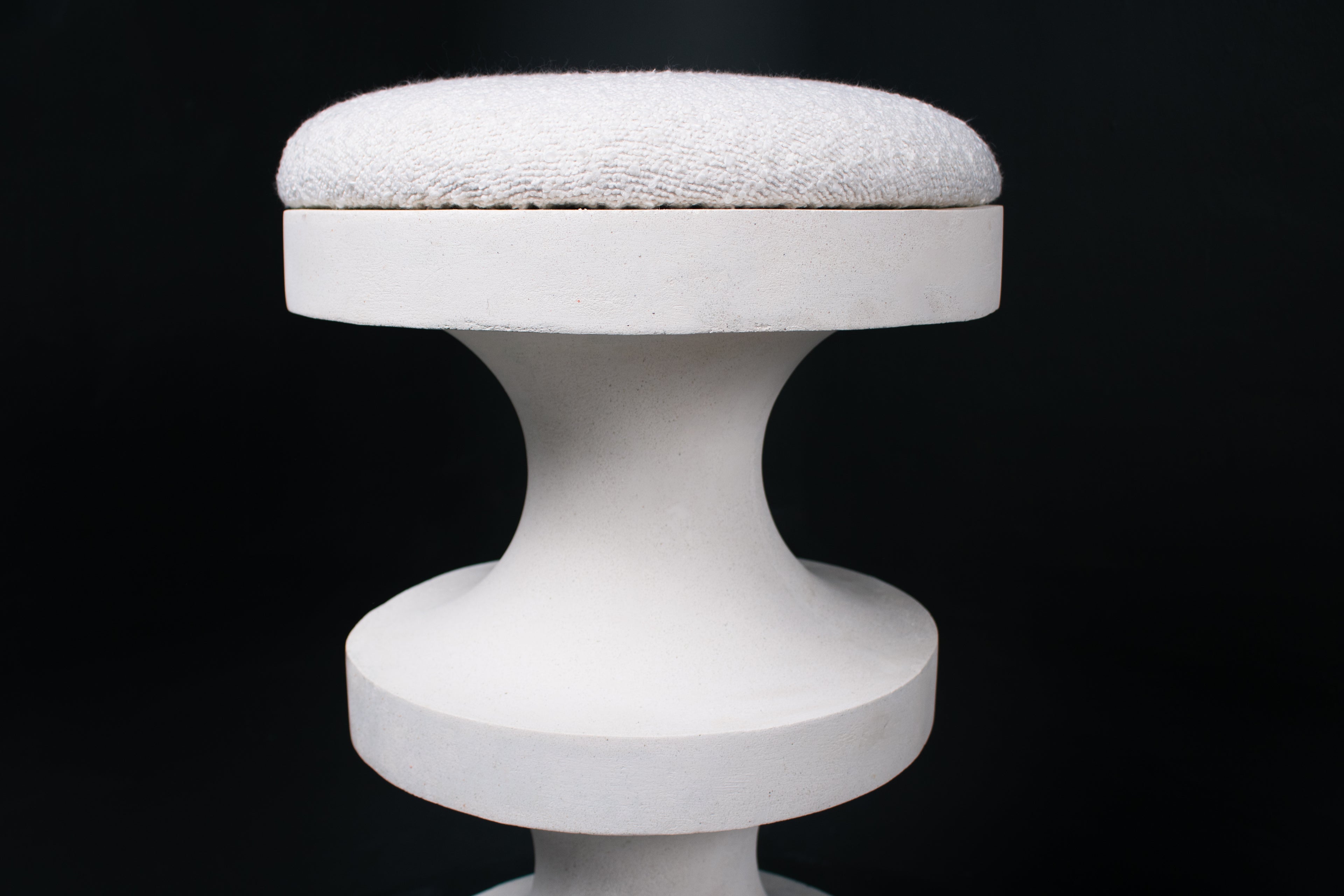The Tambour barstool in white. The sculptural pedestal base features three stacked hourglass tiers that widen and narrow in alternating rhythm, topped with a round white bouclé upholstered seat.