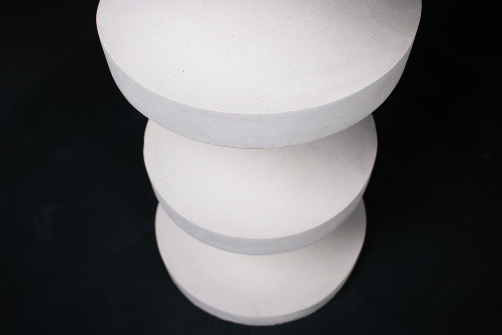 The Tambour barstool in white. The sculptural pedestal base features three stacked hourglass tiers that widen and narrow in alternating rhythm, topped with a round white bouclé upholstered seat.