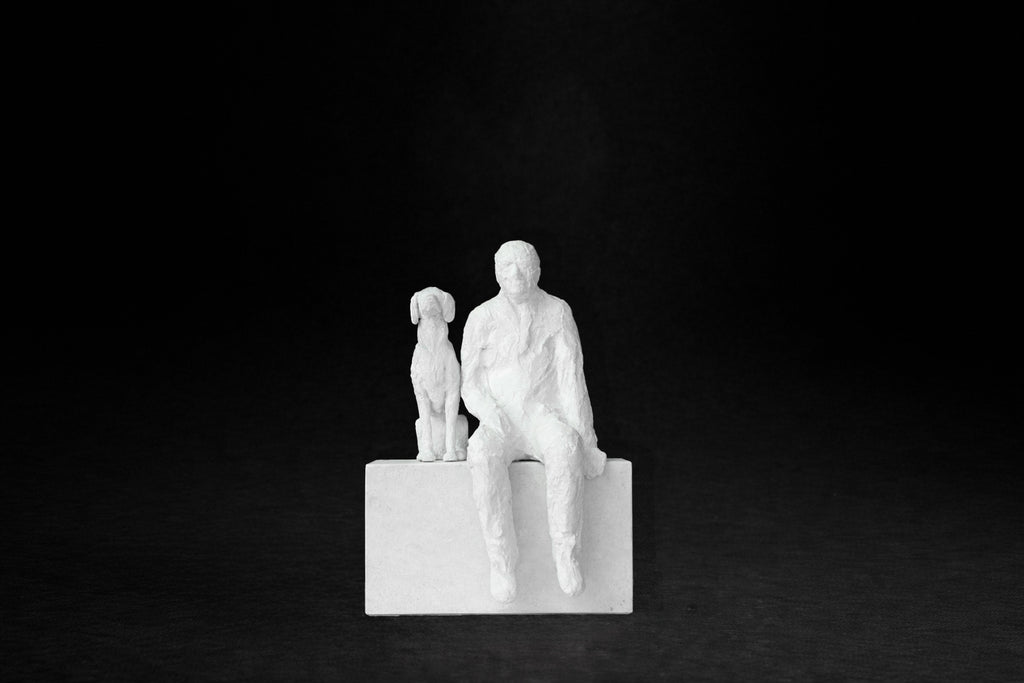 White plaster sculpture of a man sitting on a stone plinth beside his dog, both gazing outward.