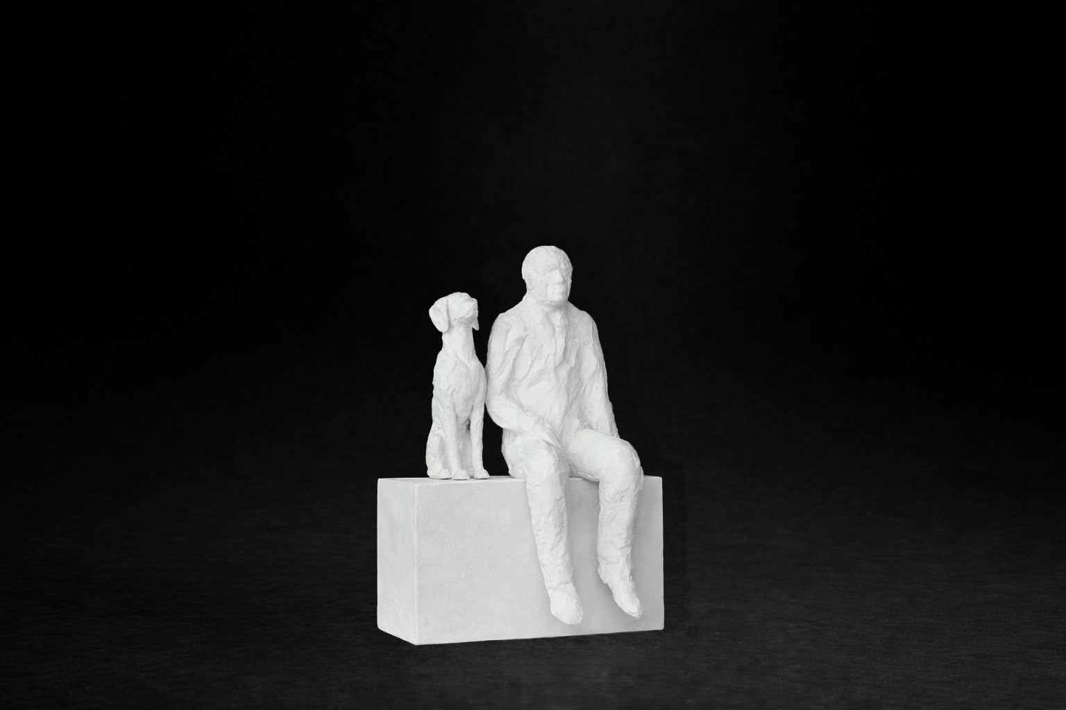 White plaster sculpture of a man sitting on a stone plinth beside his dog, both gazing outward.