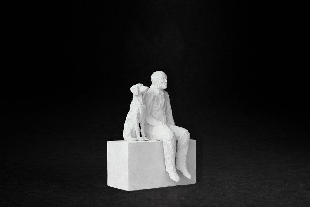 White plaster sculpture of a man sitting on a stone plinth beside his dog, both gazing outward.