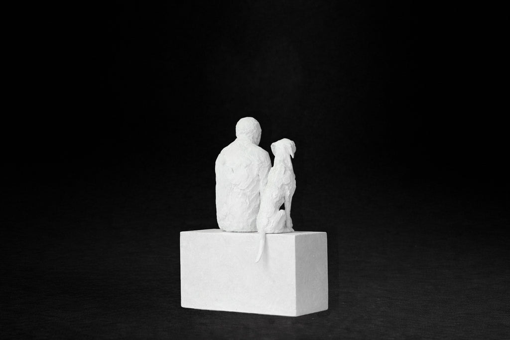 White plaster sculpture of a man sitting on a stone plinth beside his dog, both gazing outward.