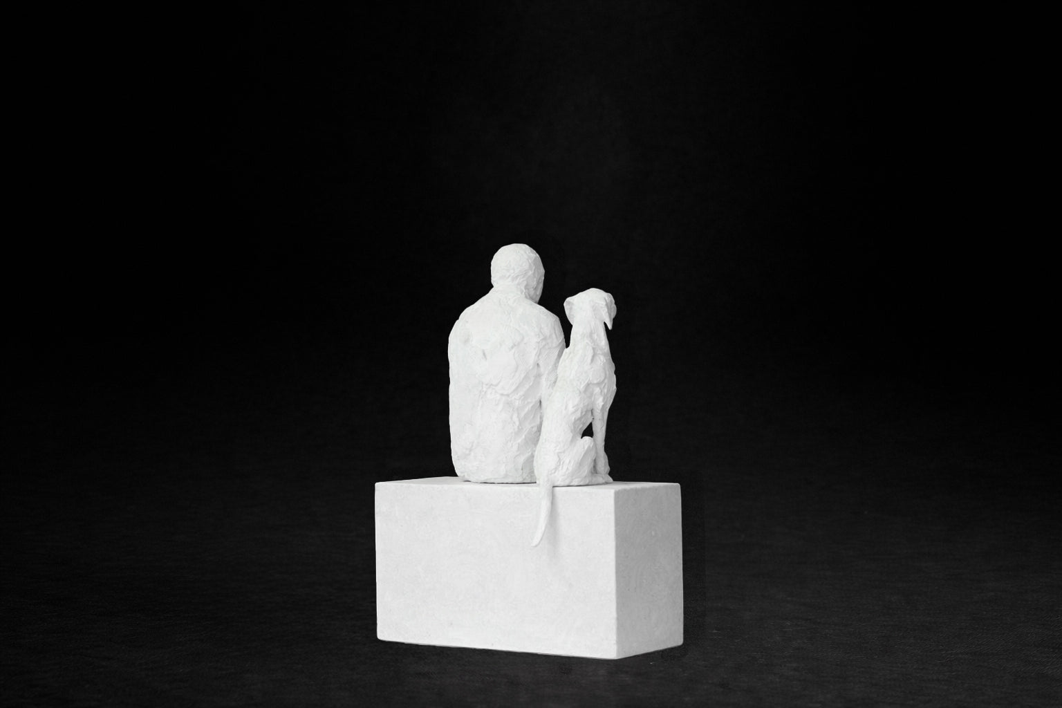 White plaster sculpture of a man sitting on a stone plinth beside his dog, both gazing outward.