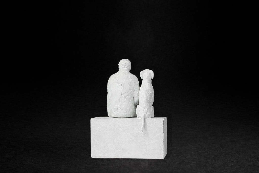 White plaster sculpture of a man sitting on a stone plinth beside his dog, both gazing outward.