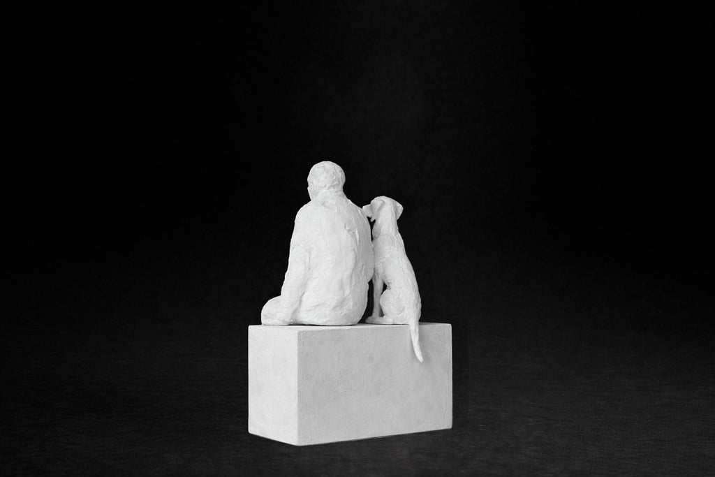 White plaster sculpture of a man sitting on a stone plinth beside his dog, both gazing outward.