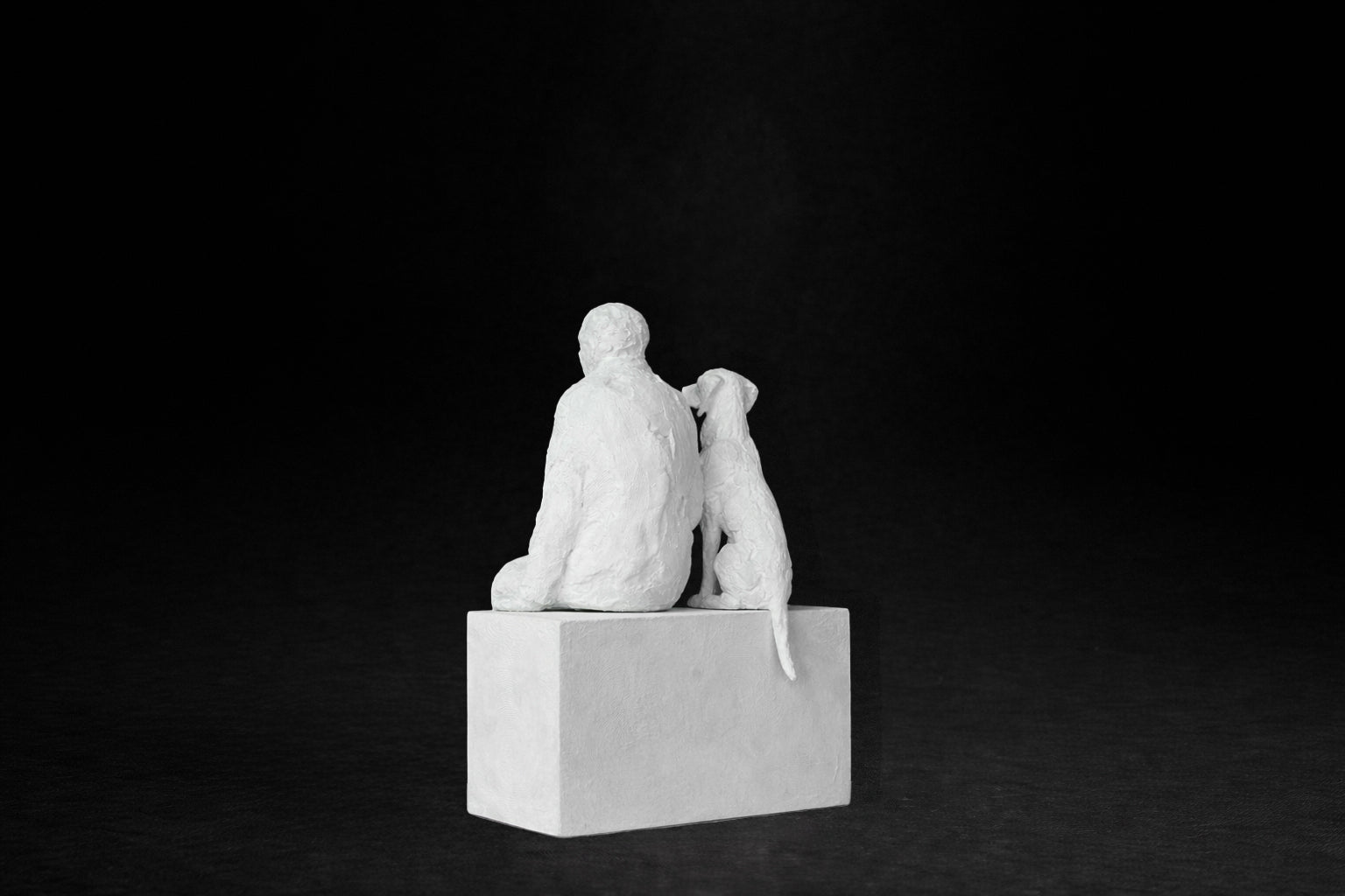White plaster sculpture of a man sitting on a stone plinth beside his dog, both gazing outward.