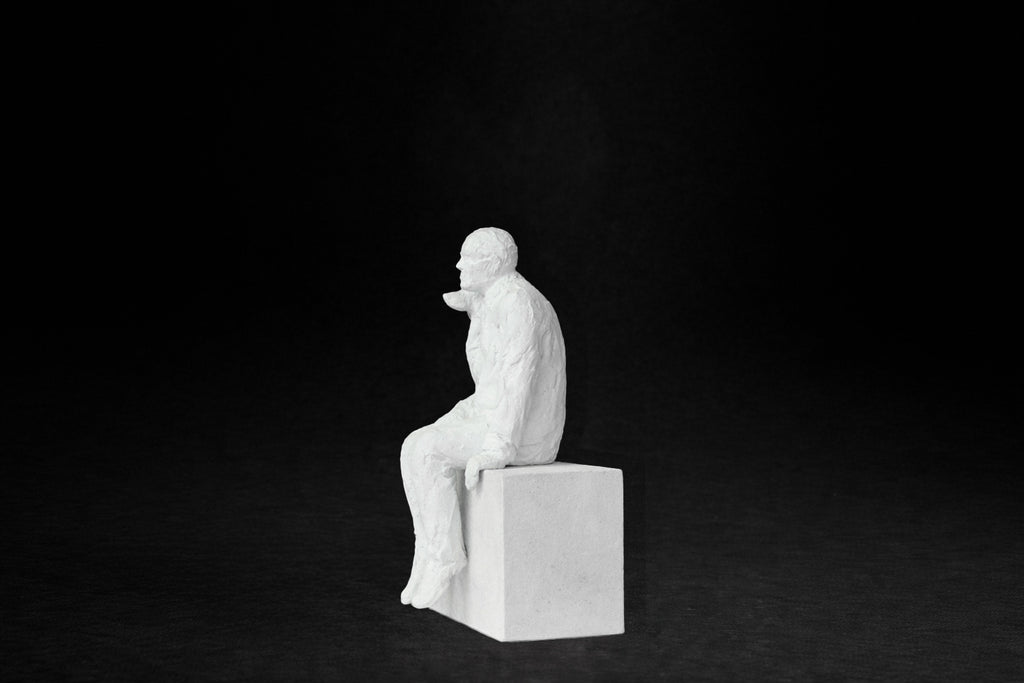 White plaster sculpture of a man sitting on a stone plinth beside his dog, both gazing outward.