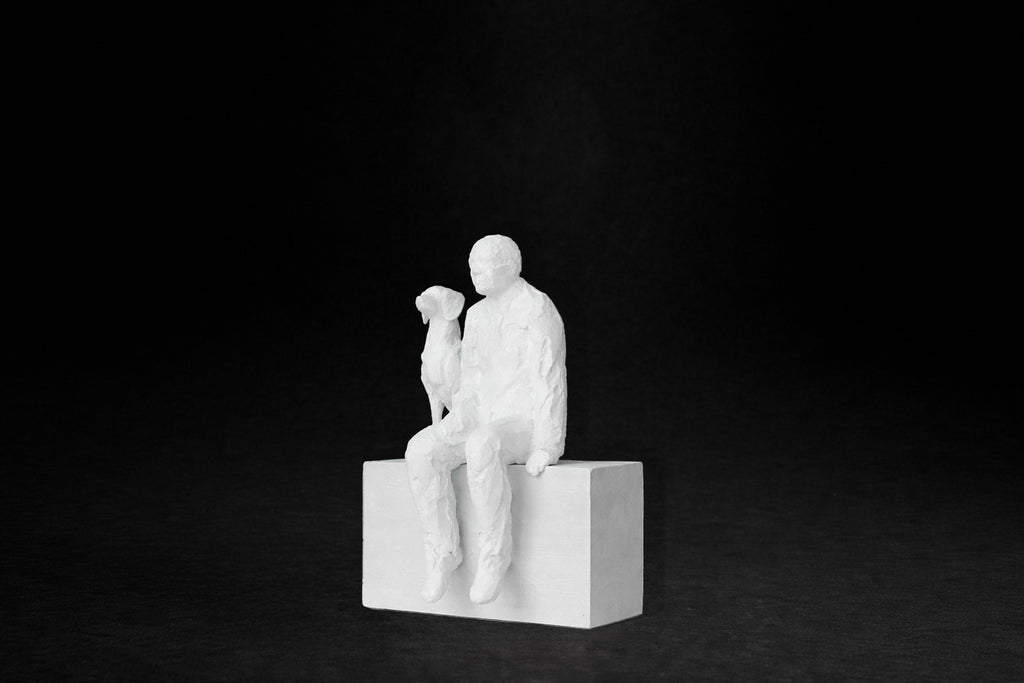 White plaster sculpture of a man sitting on a stone plinth beside his dog, both gazing outward.