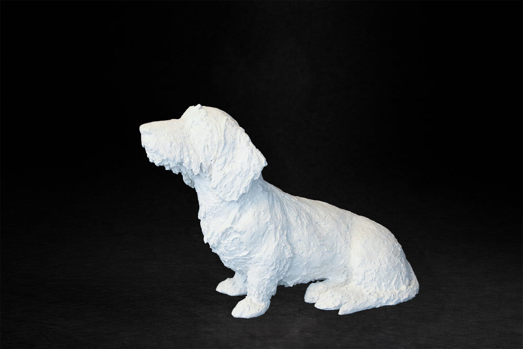 White plaster sculpture of a seated dachshund with an expressive upward gaze.
