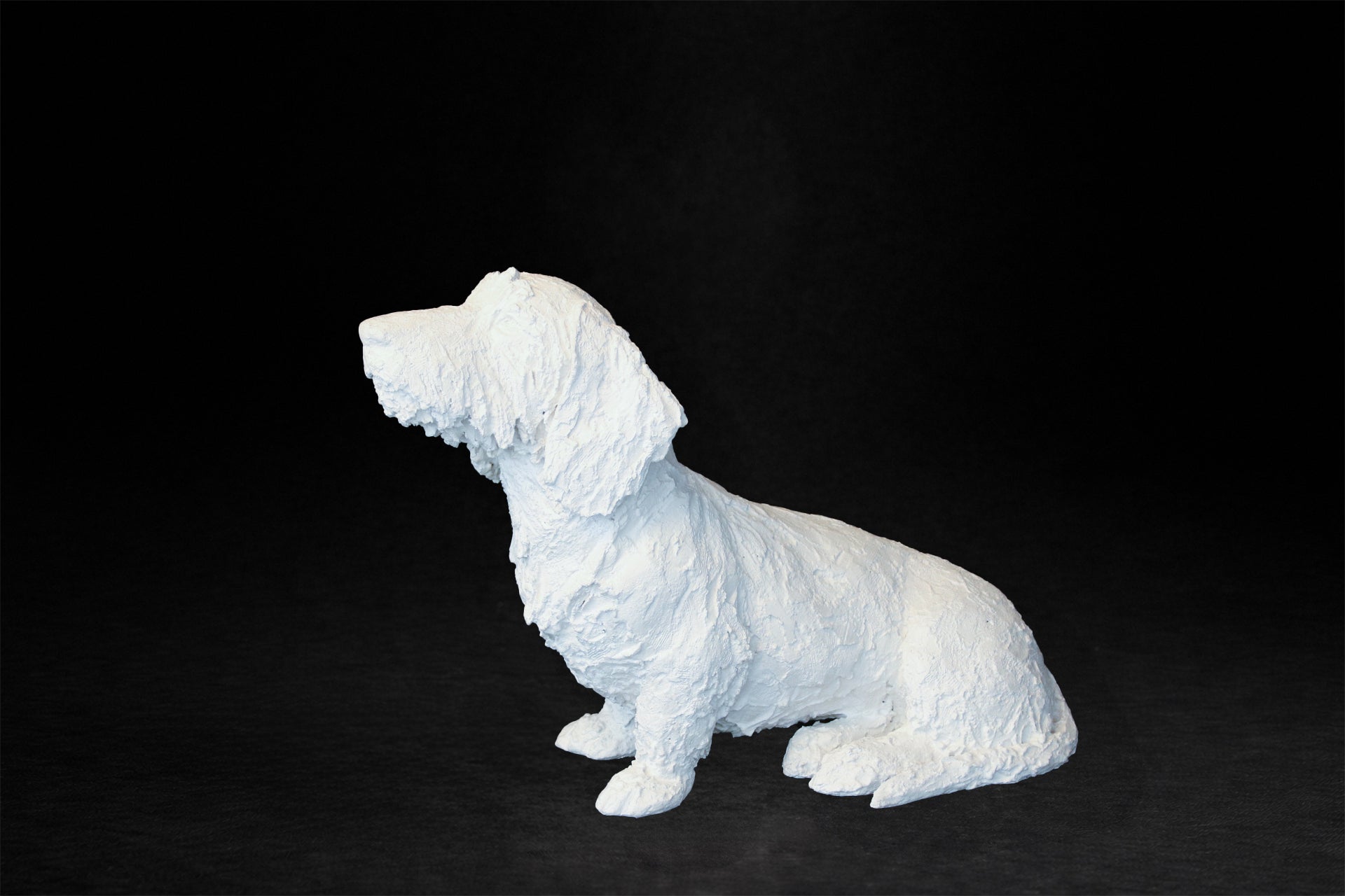 White plaster sculpture of a seated dachshund with an expressive upward gaze.