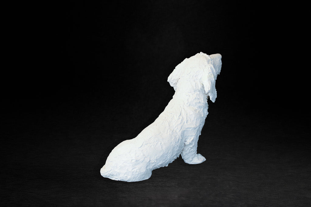 White plaster sculpture of a seated dachshund with an expressive upward gaze.