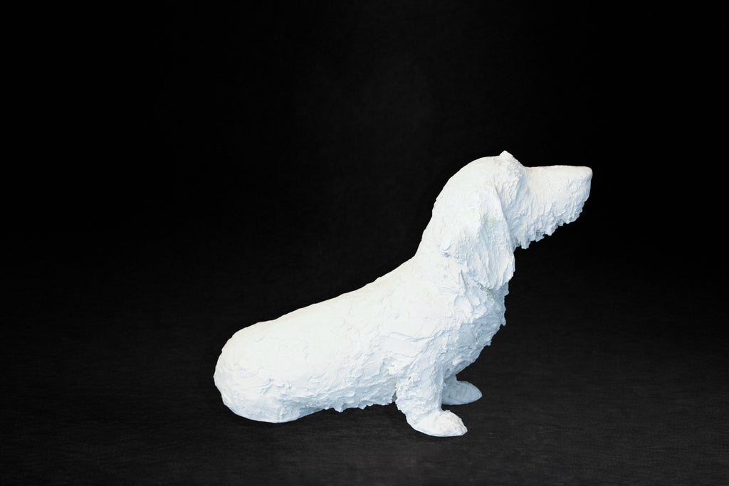 White plaster sculpture of a seated dachshund with an expressive upward gaze.