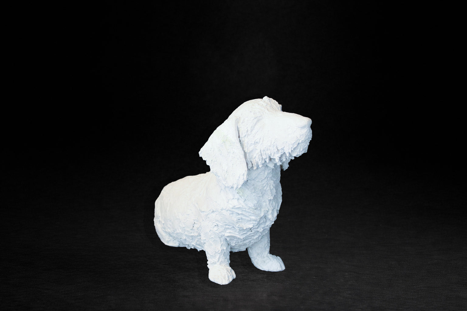 White plaster sculpture of a seated dachshund with an expressive upward gaze.